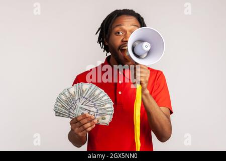 Ritratto di uomo con Dreadlocks indossare rosso casual stile T-shirt, urlando in megaphone che tiene fan di dollari, vacanze bonus e promozioni. Studio interno girato isolato su sfondo grigio. Foto Stock