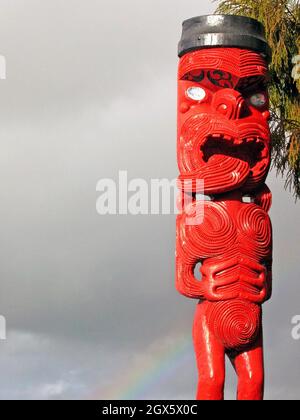 Maori Toi Whakairo, rosso e nero a tre dita, con arcobaleno in una giornata nuvolosa a Rotorua, nuova Zelanda. Le sculture in legno nella cultura Maori sono associate alla narrazione e alla spiritualità. Foto Stock