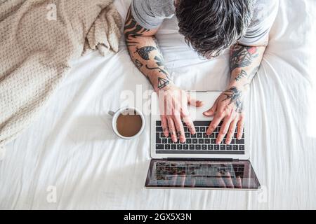 vista ritagliata dell'uomo tatuato che digita sul laptop a letto con tazza di caffè e computer portatili Foto Stock