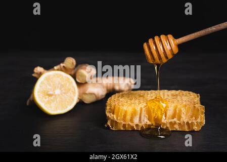 miele che gocciola da un tuffatore di legno sul nido d'ape vicino al limone sfocato e radice di zenzero isolato su nero Foto Stock