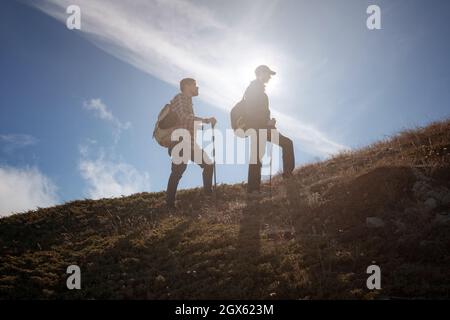 Due silhouette uomo che camminano lungo la cima della montagna con zaini da trekking si incontrano i raggi del sole e il cielo blu sfondo Foto Stock