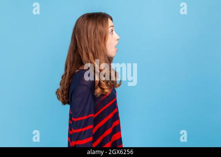 Vista laterale della donna sorpresa stupita indossando un pullover casual a righe guardando avanti con espressione scioccata, vede qualcosa di sorprendente. Studio interno girato isolato su sfondo blu. Foto Stock