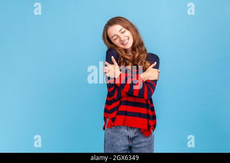 Ritratto di bella femmina in maglia casual stile a righe, in piedi abbracciandosi e sentirsi bene, apprezzando il suo aspetto, concetto di autostima. Studio interno girato isolato su sfondo blu. Foto Stock