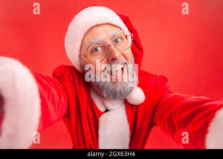 Felice uomo anziano divertente con barba grigia indossando il costume di babbo natale facendo selfie POV, guardando la macchina fotografica con sorriso ed espressione positiva. Studio interno girato isolato su sfondo rosso. Foto Stock