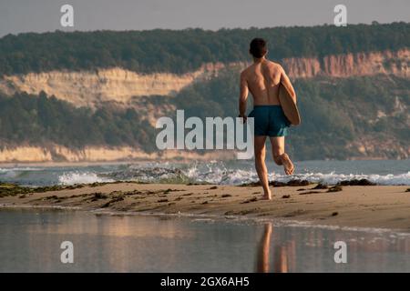 Il giovane sportivo corre con la sua skimminboard sulla spiaggia guardando l'alba Foto Stock
