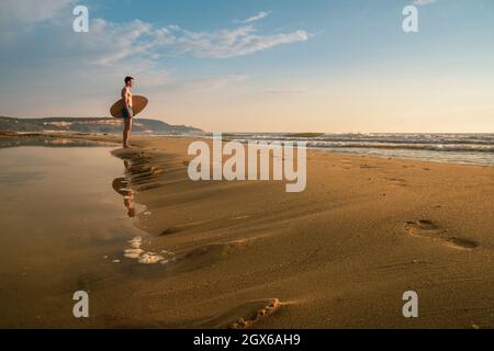 Il giovane uomo sportivo tiene la sua skimboard sulla spiaggia guardando l'alba Foto Stock