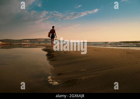 Il giovane uomo sportivo tiene la sua skimboard sulla spiaggia guardando l'alba Foto Stock