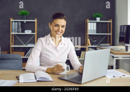 Donna d'affari felice e di successo seduta sul suo posto di lavoro con una tazza nelle sue mani. Foto Stock