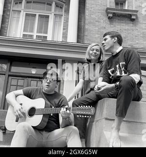 Anni '60, storico, tre studenti, uno con chitarra acustica, gli altri due canti, all'esterno del campus della Bowling Green University, Ohio, USA. Il giovane ha una chitarra goya, un marchio usato all'epoca dal co strumento musicale Hershman di New York, che li importava dalla società Levin in Svezia, rimproverandoli 'Goya', dopo l'artista spanista Francisco Goya, noto per aver presentato chitarre nei suoi dipinti. In quest'epoca, gli americani si sono presi un simpatia per la chitarra Goya d'epoca, che si è rivelata popolare soprattutto con i musicisti folkloristici. Foto Stock