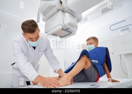 Medico professionista in uniforme bianca facendo visita medica prima della diagnostica a ultrasuoni. Ragazzo seduto in clinica e check in gamba dopo il dolore. Concetto di medicina dei bambini. Foto Stock