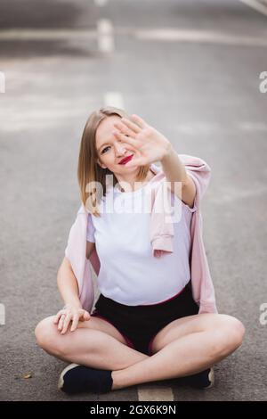 Giovane donna felice in t-shirt bianca si siede in mezzo alla strada asfaltata e mostra fermarsi con la mano Foto Stock