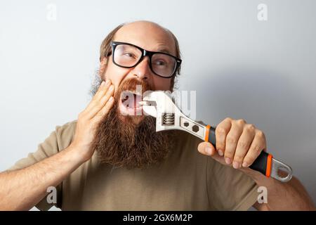 Maschio adulto che tira fuori il dente cattivo da solo, toccando guancia con mano, urlando da dolore terribile, uomo calvo ported indossando T-shirt e occhiali. Studio interno girato isolato su sfondo grigio. Foto Stock