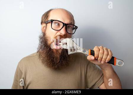 Divertente maschio che tira fuori il dente con la chiave, avendo espressione stolish, guarda via con gli occhi incrociati, uomo calvo bearded che indossa la T-shirt e gli occhiali. Studio interno girato isolato su sfondo grigio. Foto Stock