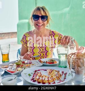 una giovane donna con occhiali da sole e capelli biondi pinned up è seduta a un tavolo riccamente impostato con un pasto con cibo e bevande all'aperto su una terrazza. Foto Stock