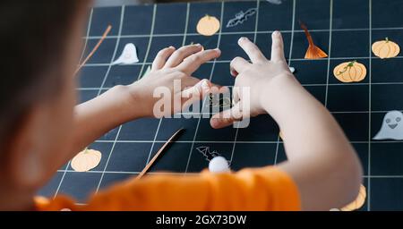 Cute bambino che crea Halloween-tema gioco da tavolo. Ragazzo incollando elementi di gioco tagliati su carta sul campo da gioco nero. Idee fai da te per i bambini Foto Stock