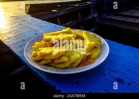Un piatto bianco contiene un ananas a fette su un sedile in legno blu durante l'alba nel mercato galleggiante del Delta del Mekong. Foto Stock