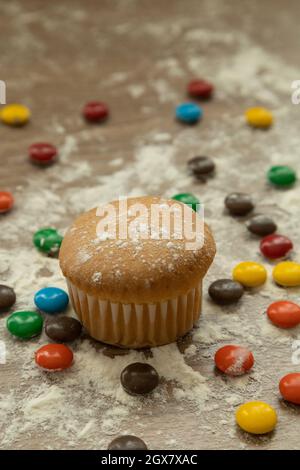 muffin dolci cotti accanto a diversi cioccolatini colorati, spolverati con zucchero sulla parte superiore, su un tavolo di legno, deliziosi dessert, cibo in studio Foto Stock