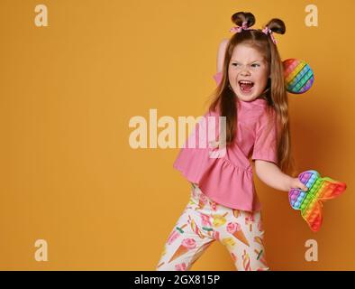 Frolic, urlando, capretto viziato in vestiti colorati che vanno gettare due giocattoli sensoriali di colore dell'arcobaleno - pop rotondo Foto Stock