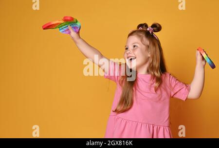 Felice ragazza rossa capretto in maglia rosa gioca con i nuovi giocattoli sensoriali del colore dell'arcobaleno - forma della farfalla e pop rotondo esso Foto Stock