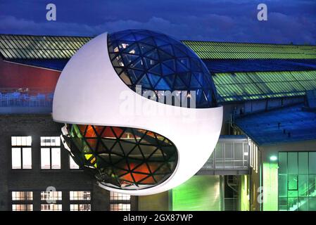 29 settembre 2021, Sassonia, Lipsia: Dopo il tramonto, la sfera illuminata Niemeyer sorge dal cielo blu sopra gli edifici del costruttore di gru Kirow e del costruttore di tram HeiterBlick sui terreni di 'Techne Sphere Leipzig' durante una serata di menu. Completata nell'estate del 2020, la sfera di cemento e vetro di 12 metri progettata dall'architetto brasiliano Oscar Niemeyer (1907-2012), ospita ora i suoi primi eventi e celebrazioni dopo la lunga pausa di Corona. La struttura a cupola con i suoi tre livelli e circa 150 triangoli di vetro colorato in cima ad un edificio industriale in mattoni è l'ultima completa Foto Stock