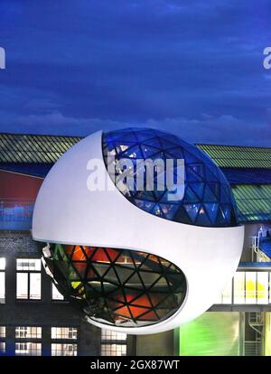 29 settembre 2021, Sassonia, Lipsia: Dopo il tramonto, la sfera illuminata Niemeyer sorge dal cielo blu sopra gli edifici del costruttore di gru Kirow e del costruttore di tram HeiterBlick sui terreni di 'Techne Sphere Leipzig' durante una serata di menu. Completata nell'estate del 2020, la sfera di cemento e vetro di 12 metri progettata dall'architetto brasiliano Oscar Niemeyer (1907-2012), ospita ora i suoi primi eventi e celebrazioni dopo la lunga pausa di Corona. La struttura a cupola con i suoi tre livelli e circa 150 triangoli di vetro colorato in cima ad un edificio industriale in mattoni è l'ultima completa Foto Stock