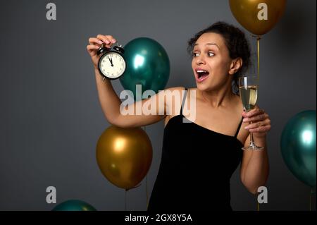 Bella donna da corsa mista in abito di velluto nero sera in posa con sveglia e bicchiere di vino frizzante contro lucido verde metallico e dorato a. Foto Stock