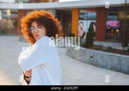Donna d'affari curly che tiene il laptop e sorprende gesturing fuori. Uomini d'affari in posa sotto il sole. Foto Stock