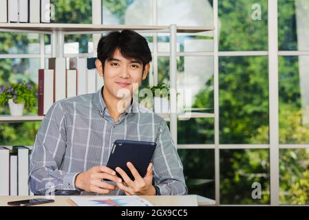 Giovane uomo d'affari asiatico seduto in ufficio sorride e lavora su un nuovo tablet, il giovane e sicuro che usa il touchpad checkin Foto Stock