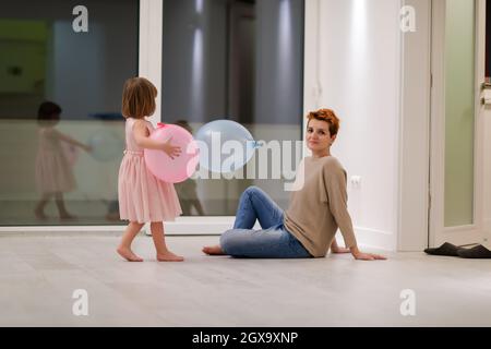 la famiglia felice che trascorre il tempo insieme giovane madre redhead e figlia piccola cute che si divertono mentre gioca con i palloncini vicino alla finestra su bello Foto Stock