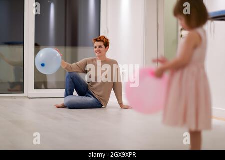 la famiglia felice che trascorre il tempo insieme giovane madre redhead e figlia piccola cute che si divertono mentre gioca con i palloncini vicino alla finestra su bello Foto Stock