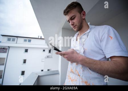 Pittore che fa la chiamata telefonica mentre si alza sul balcone durante la pausa dal decoratore di lavoro Usando il telefono mobile in piedi Fuori Foto Stock