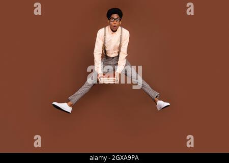 Vista a tutta lunghezza del corpo di attraente ragazzo divertente stupito che salta tenendo libro accademico isolato su sfondo di colore marrone. Foto Stock