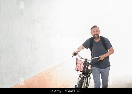 Un giovane sorridente elegante tanga barbuto uomo con uno zaino spingendo una bicicletta retrò mentre si cammina sulla strada Foto Stock