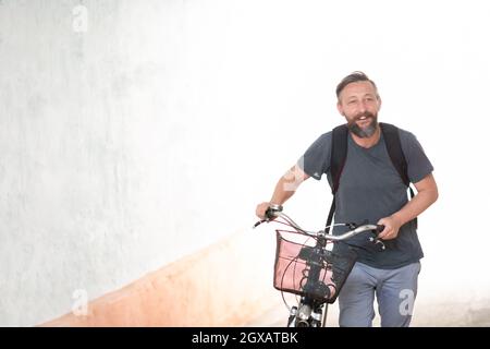 Un giovane sorridente elegante tanga barbuto uomo con uno zaino spingendo una bicicletta retrò mentre si cammina sulla strada Foto Stock