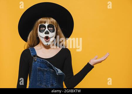 Felice Halloween concetto. Ritratto di bambino scioccato ragazza con trucco spooky indossa grande cappello nero mostrando spazio vuoto copia sul palmo aperto della mano Foto Stock
