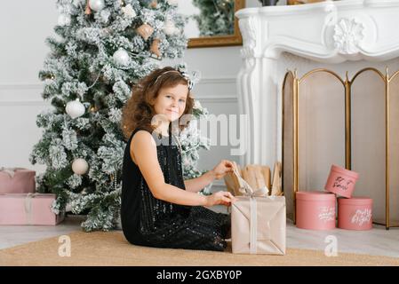 Una bambina di sette anni in abito nero con paillettes si siede vicino al camino all'albero di Natale e apre un regalo di Capodanno Foto Stock
