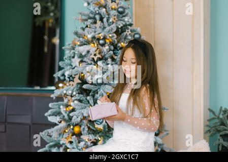 Carina bambina in un abito bianco è seduta sul divano e si apre una scatola regalo sullo sfondo di un albero di Natale con decorazioni in oro. Il concetto Foto Stock