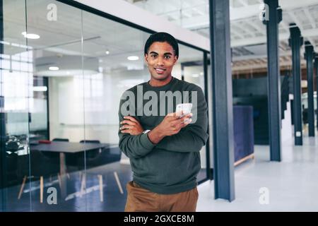 Giovane uomo d'affari creativo che tiene uno smartphone in un ufficio moderno. Felice giovane uomo d'affari che guarda la fotocamera mentre si alza in piedi da solo in un posto di lavoro. Foto Stock