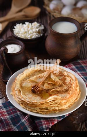 Frittelle fatte in casa con miele accatastato in una pila, su un tavolo di legno con una tazza di latte, una pentola di panna acida e uova in un cestino. Tradizionale slavo Foto Stock