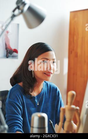 Sorridente giovane donna asiatica in maglione blu seduta in ufficio e lavorando al design grafico Foto Stock
