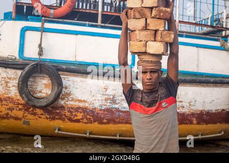 INDIA, BANGLADESH - 8 DICEMBRE 2015: Giovane etnia maschio in abiti sporchi camminare portando pietre di mattoni sopra testa vicino al fiume con le barche che guardano è venuto Foto Stock
