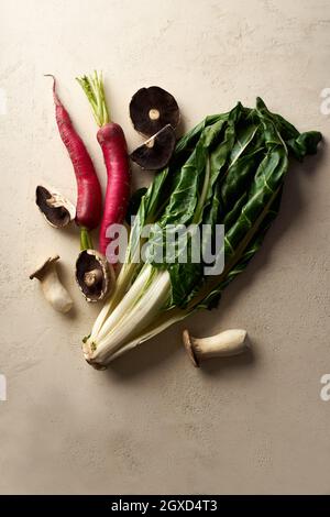 Verdure e funghi biologici su sfondo beige. Vista dall'alto con green sani e daikon rosso invernale. Nuovi ingredienti nella routine alimentare sana. Foto Stock