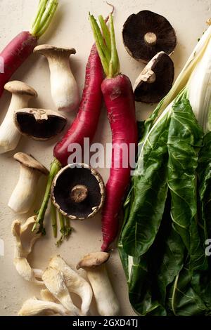 Verdure e funghi biologici su sfondo beige. Primo piano con green sani e daikon rosso invernale. Nuovi ingredienti nella routine alimentare sana. Foto Stock