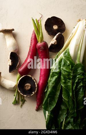 Verdure e funghi biologici su sfondo beige. Vista dall'alto con green sani e daikon rosso invernale. Nuovi ingredienti nella routine alimentare sana. Foto Stock