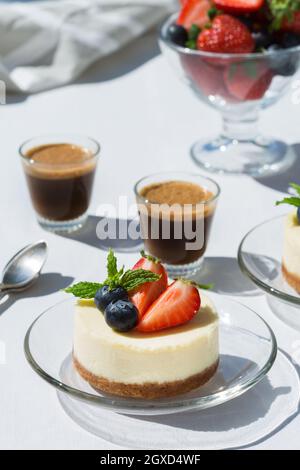 Gustosi cheesecake con fragole fresche e mirtilli sotto le foglie di menta contro shot di caffè espresso sul tavolo in caffetteria Foto Stock