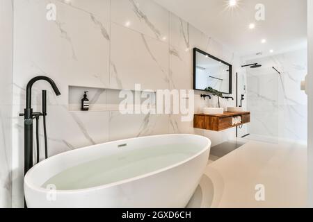 Acqua pura in vasca da bagno ovale contro il lavabo sotto lo specchio e lampade lucenti nella stanza da bagno contemporanea in casa Foto Stock