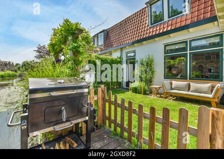 Barbecue chiuso posto vicino recinzione in legno sulla terrazza con erba verde e piante vicino cottage moderno in estate Foto Stock