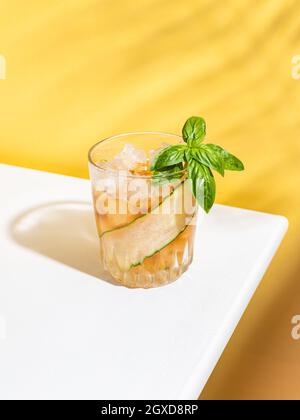 Alto angolo di bicchiere di cocktail freddo con fette di cetriolo e foglie di basilico servito su tavola bianca su sfondo giallo Foto Stock