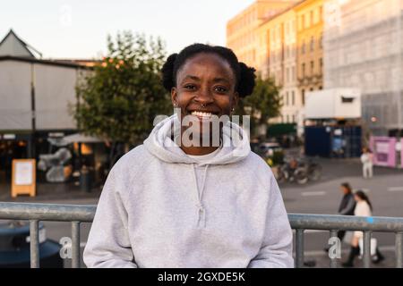 Felicissima femmina androgina afroamericana che sorride ampiamente mentre si trova sulla recinzione della strada della città guardando la macchina fotografica Foto Stock