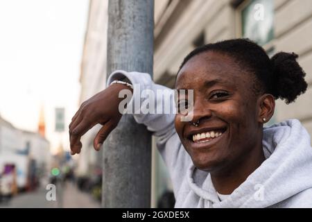 Felicissima femmina androgina afroamericana sorridente ampiamente mentre si appoggia sul pilastro di metallo e guarda la macchina fotografica Foto Stock